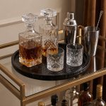 Black marble round tray on a gold bar cart holding a whiskey decanter, crystal glasses and cocktail shaker